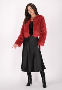 Giacca corta in pelliccia sintetica rossa con maniche lunghe, texture stratificata. Una gonna midi in satin nera e stivali con tacco al ginocchio completano l'outfit.