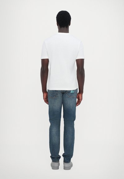 T-shirt blanc à manches courtes, jeans en denim bleu et chaussures grises. Les jeans ont une étiquette verte sur la poche arrière. Vue de dos.