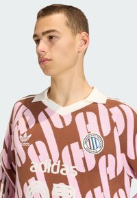 Bruin en roze gebreide jersey met patroon en een witte kraag. Heeft het Adidas-logo op de borst en een ronde badge met gekleurde strepen.