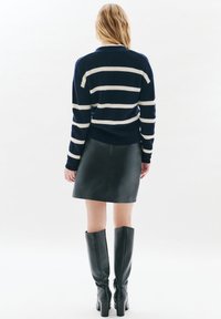 Kobieta z długimi blond włosami ubrana w granatowy sweter w białe paski, czarną skórzaną mini spódnicę i czarne kozaki do kolan, stoi tyłem.