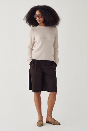 Kobieta o bujnych kręconych włosach, nosząca ciemne okulary przeciwsłoneczne, beżowy sweter, czarne krótkie spodenki za kolano oraz beżowe wsuwane buty, stojąca z rękami w kieszeniach.