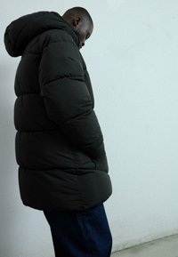 Svart pufferjacka med huva, med en quiltad design, tjock stoppning och en sidoficka, i kombination med mörka denimjeans.