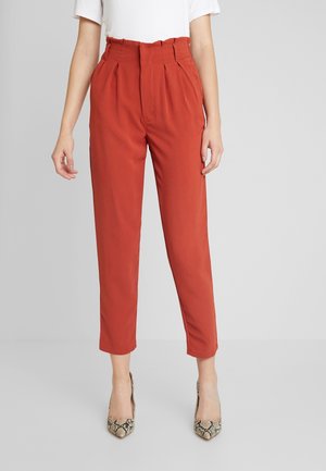 Femme portant un pantalon fuselé taille haute couleur rouille avec des plis, associé à des talons pointus imprimé serpent et un top blanc à manches courtes.