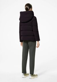 Giacca a vento violacea scura con un ampio cappuccio e trapuntatura orizzontale, abbinata a pantaloni verdi e sneakers olive. Texture liscia, design moderno.