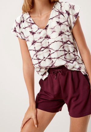 Frau trägt eine weiß-lila gemusterte V-Ausschnitt-Bluse mit kurzen Ärmeln und einfarbige bordeauxrote Shorts mit Kordelzug.
