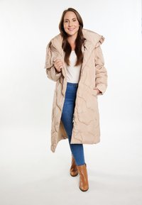 Cappotto imbottito beige con design a trapunte, grande cappuccio e chiusura con zipper, abbinato a jeans blu e stivali alla caviglia marroni con tacco.