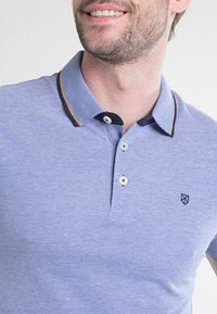 Jasnoniebieska koszulka polo o teksturowanym splotzie, z kontrastującym czarnym kołnierzykiem i żółtymi wykończeniami oraz małym logo na piersi. Trzy guziki.