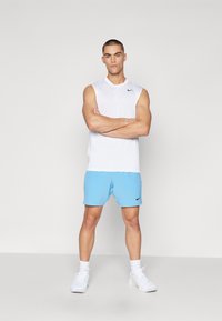 Valkoinen hihaton urheilupaita, jossa on musta Nike-logo, yhdistettynä vaaleansinisiin shortseihin ja valkoisiin lenkkareihin, neutraalilla taustalla.
