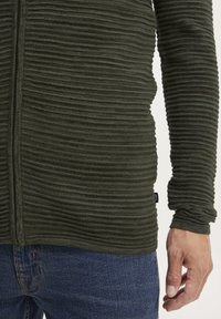 Grønstribet langærmet top med en frontlynlås, udstyret med en ribbet tekstur, parret med denim jeans og et synligt mærke på venstre side.
