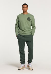 Felpa verde con un motivo esagonale nero sul petto, abbinata a pantaloni della tuta verde scuro e scarpe da ginnastica multicolori.
