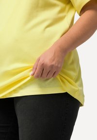 Žluté krátké tričko s rukávy vyrobené z měkké bavlny, s volným střihem a plisováním u lemu. Nosené přes tmavé džíny.