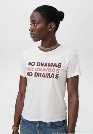 Une femme à la peau foncée et aux cheveux relevés en chignon porte une chemise blanche sur laquelle est inscrit "PAS DE DRAMAS" trois fois dans différentes nuances de rouge et de rose.
