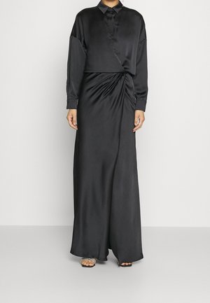 Kvinde iført en lang sort satin kjole med lange ærmer, en krave og subtil drapering ved taljen, kombineret med åbne strop sandaletter.