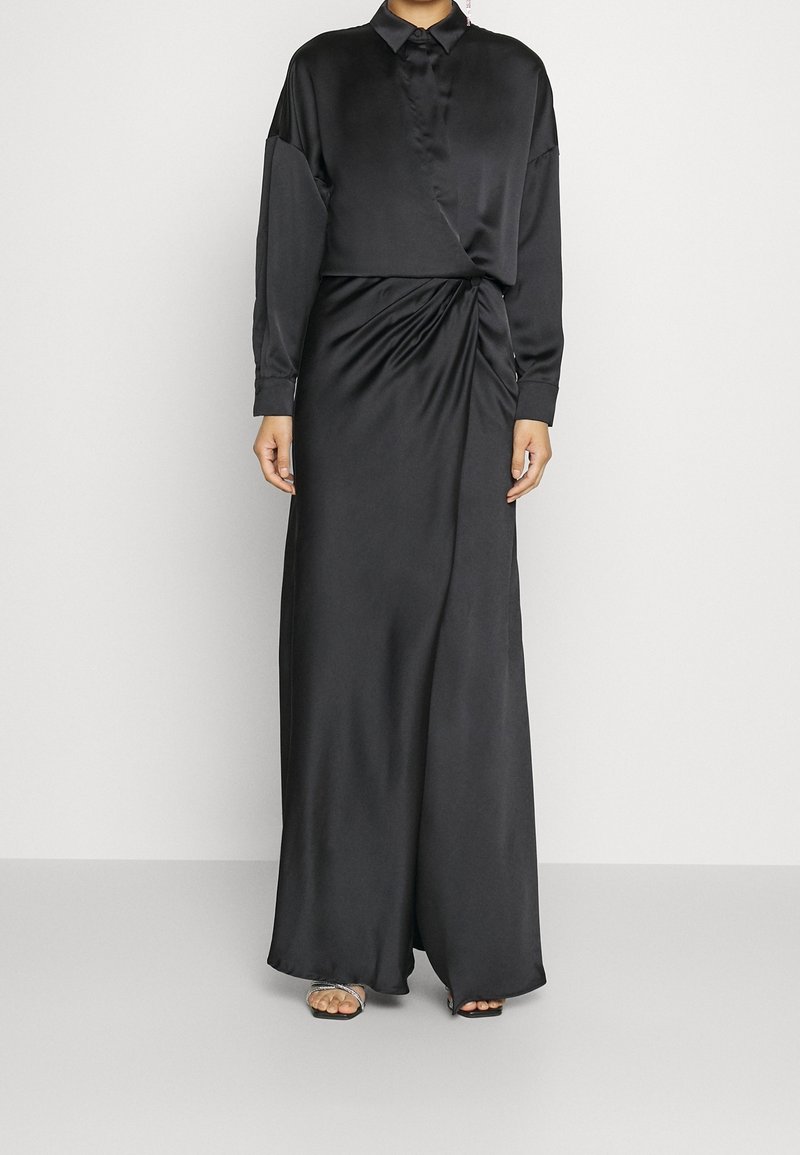 Femme portant une longue robe en satin noir à manches longues, col et un drapé subtil à la taille, associée à des talons ouverts à brides.