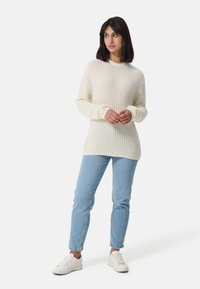 Sweter dzianinowy w kolorze kremowym, z prążkowaną teksturą i wysokim kołnierzem. W zestawie z jasnoniebieskimi dżinsami i białymi sneakersami.