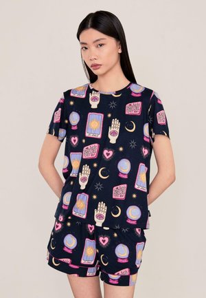Mujer con cabello negro liso que lleva una camisa y pantalones cortos azul oscuro a juego con símbolos coloridos de adivinación y motivos celestiales.