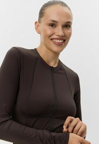 Hnedý dlhý rukáv top vyrobený z elastického materiálu, s predným zipsom a viditeľnými švovými akcentmi. Jednoduchý dizajn s tesným strihom.