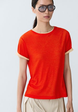 Femme portant une jupe beige portefeuille, un top orange vif à manches courtes avec bordure beige et des lunettes de soleil surdimensionnées, debout devant un fond clair uni.