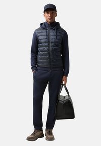 Marineblå polstret vest over en mørk træningsdragt, parret med brune sneakers. Personen holder en sort, struktureret duffelbag og bærer en matchende kasket.