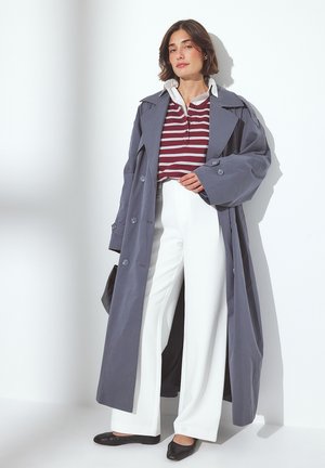 Nainen, jolla on päällään pitkä harmaa trench coat, burgundy- ja valkoisraidallinen polo-paita, valkoiset leveälahkeiset housut ja mustat ballerinat, nojautuu valkoista seinää vasten.