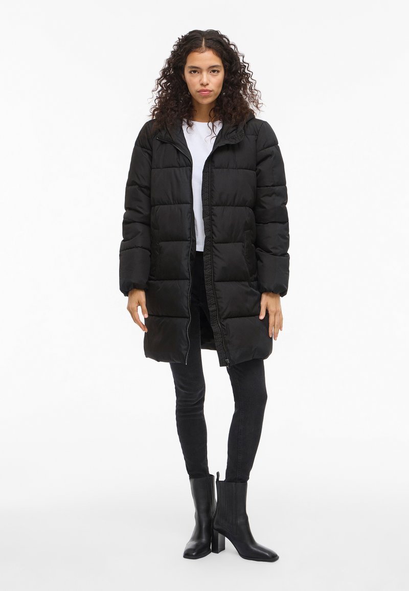 Crna puffer jakna s kapuljačom, s prednjim patentnim zatvaračem i velikim prošivenim kvadratima. Uparena s crnim uskim trapericama i crnim gležnjačama s potpeticom.