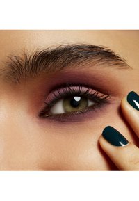 Close-up van een oog met een groene iris, gemengde paarse oogschaduw, lange wimpers en donkere wenkbrauwen. Een hand met teal nagellak raakt de wang aan.