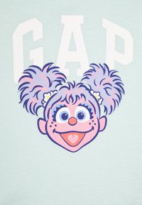 Gaiši mentola krāsas T-krekls ar karikatūras varoni ar purpura un rozā pūkainiem matiem, lielām acīm un apaļu degunu, ar vārdu "GAP" treknajās baltajās burtos.