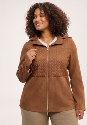 Smilende kvinde med krøllet hår iført brun quiltet lynlåsjakke og beige bukser, stående mod en ensfarvet lys baggrund.