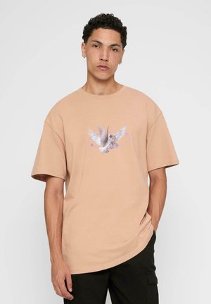 Oversized peach t-shirt made of cotton, featuring a centered graphic of a bird with wings spread and textual accents in purple.