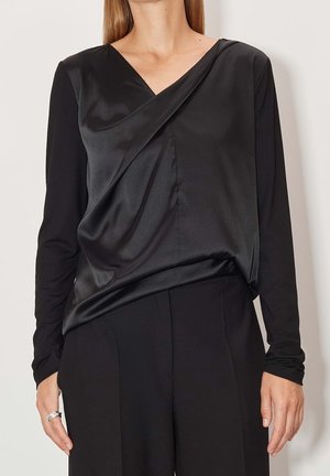 Žena v černé blůze s dlouhými rukávy se saténovým předním dílem a černých pouzdrových kalhotách, stojící před jednobarevným pozadím.