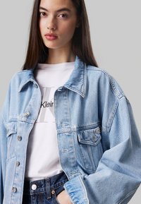 Lichtblauwe denimjas met een ontspannen pasvorm, voorzien van zilverkleurige hardware, borstzakken en een zachte textuur, gelaagd over een witte graphic tee.