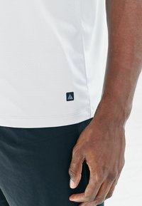 Weißes, strukturiertes Sport-Shirt mit einem schwarzen Logosticker am Saum, kombiniert mit dunklen Hosen, die die Details des Stoffes und die Passform hervorheben.