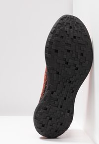 Červeno-černá teniska s texturovanou podrážkou se vzorem mřížky z gumy. Vrchní část je z pleteného materiálu s oranžovými skvrnami.