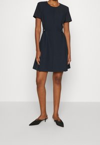 Marinblå klänning med korta ärmar, utsvängd kjol, framstygn och dekorrande knappar på sidan. Matchas med svarta spetsiga mules.