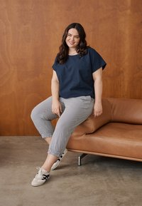Femme aux cheveux foncés portant un haut bleu marine, un pantalon rayé et des baskets, assise au bord d'un canapé en cuir marron contre un mur en bois.