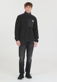 Veste en polaire noire avec un col montant, poche poitrine zippée en tissu sombre et deux poches latérales. Associée à un jean gris et des chaussures noires.