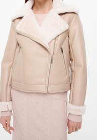 Giacca in finta pelle beige con un morbido collare e polsini in pelliccia di pecora crema, dotata di tasche anteriori con zip e design aderente.