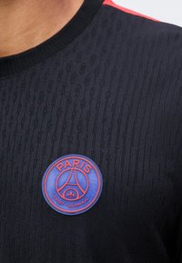 Κοντινή λήψη μαύρου υφασμάτινου πουκαμίσου με στρογγυλή μπλε και κόκκινη πατς λογότυπου του Παρί Σεν Ζερμέν στο στήθος.