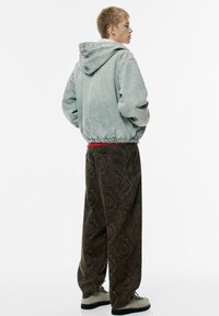 Persoană tânără stând de profil, îmbrăcată cu o jachetă cu glugă de culoare gri deschis, pantaloni largi cu model de culoare maro și pantofi texturați de culoare bej.