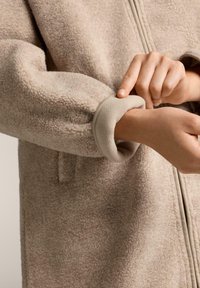 Bež flis jakna z visokim ovratnikom, zadrgo in elastičnimi manšetami. Tkanina ima mehko, teksturirano površino.