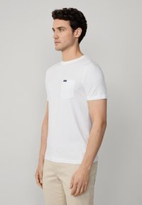 Camiseta blanca de algodón con cuello redondo, mangas cortas y un bolsillo en el pecho. Presenta un pequeño logo texturizado en el bolsillo. Textura suave.