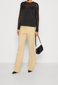 Femme portant un haut en dentelle noire à manches longues, un pantalon beige ajusté, des chaussures noires pointues, tenant un sac à main noir par la lanière.