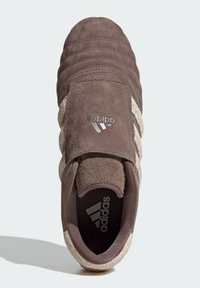 Brown suede sneaker featuring three beige stripes, textured upper, and a round toe. Adidas logo appears on the side and inside the shoe.