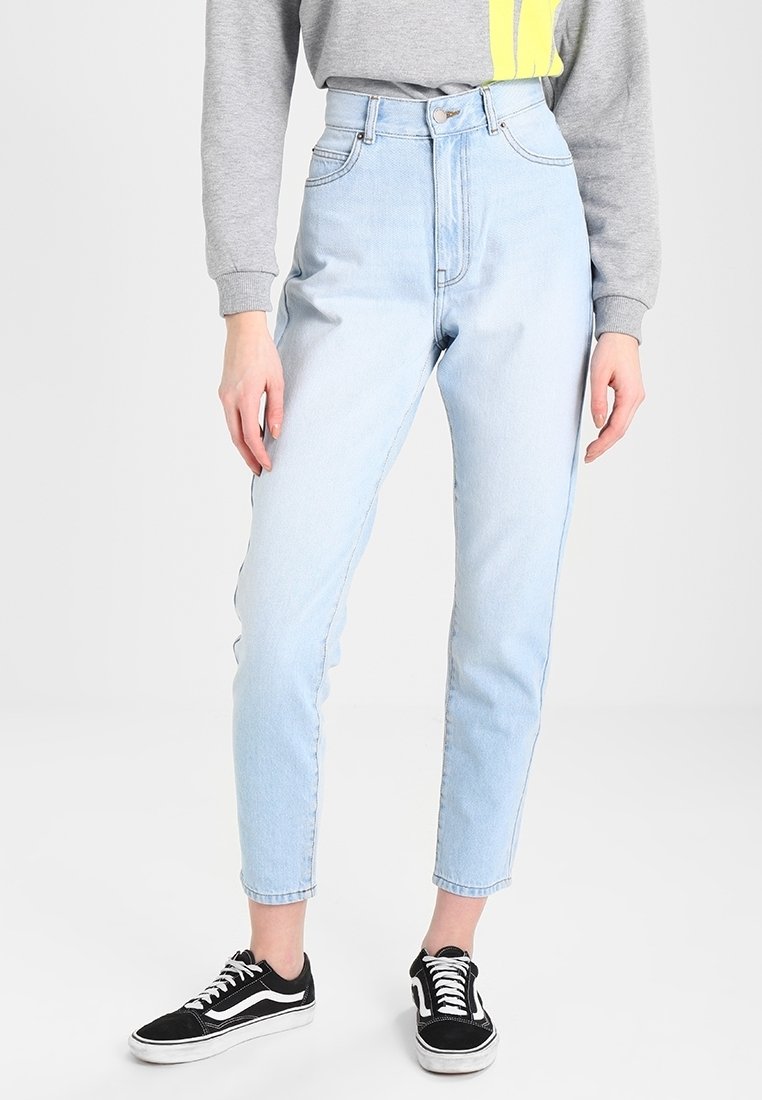 Personne portant un jean taille haute bleu clair, un sweat gris avec une bande jaune, et des baskets basses noires avec des lacets et semelles blancs.