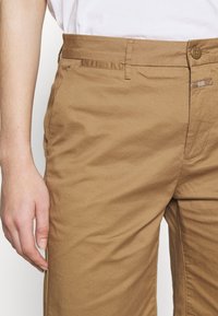 Jasnobrązowe bawełniane spodnie z bocznymi kieszeniami, zapięciem na guziki i widocznymi detalami szwów. Tkanina ma gładką, lekko teksturowaną powierzchnię.