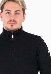 Giovane uomo con capelli castano chiaro e biondi che indossa un maglione nero con zip, rivolto in avanti su uno sfondo bianco.