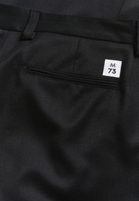 Čierne nohavice z textúrovanej tkaniny, so zadnou vreckom s čiernym prešívaním a bielou nálepkou označenou "M 73."