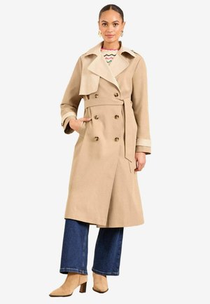 Beige Trenchcoat mit doppelt geknöpften Leisten, weitem Kragen und Gürtel. Kombiniert mit blauen weit geschnittenen Jeans und beigen Stiefeletten mit Absatz.