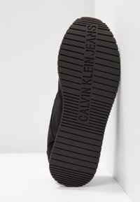 Semelle d'une chaussure noire Calvin Klein Jeans avec des rainures horizontales et le nom de la marque embossé verticalement au centre.