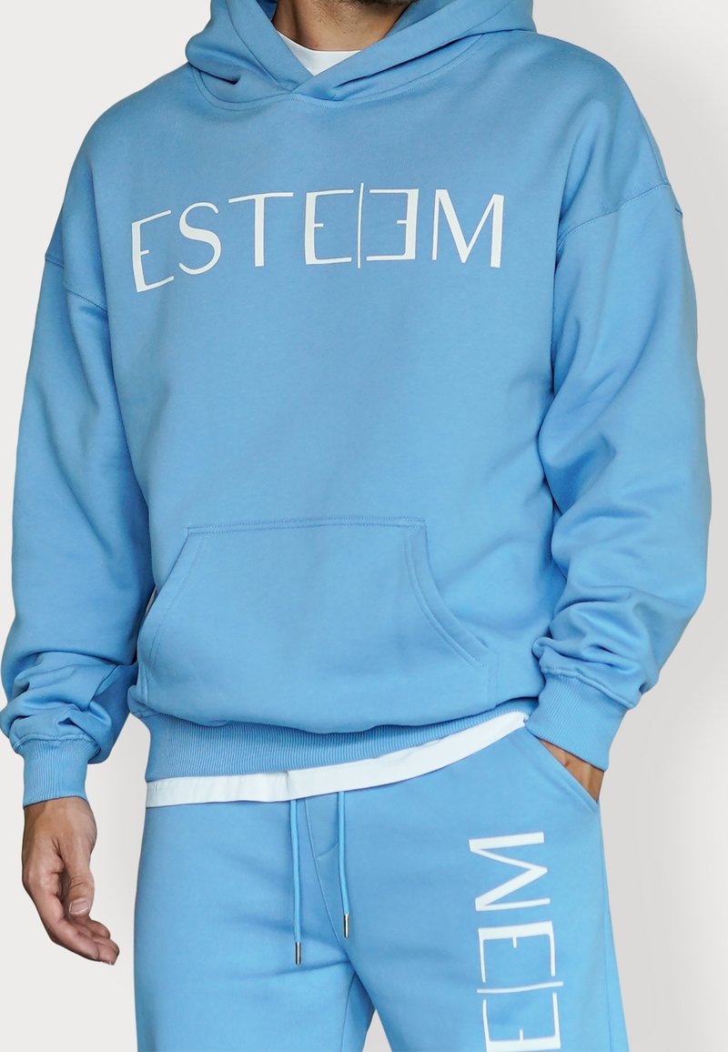 Homme portant un sweat à capuche bleu clair et un pantalon de survêtement assorti avec le texte blanc « ESTEEM » sur la poitrine et la jambe, debout devant un fond uni.